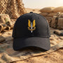 A black baseball cap with a gold and silver military-style SAS Who Dares Wins insignia on the front, resting on a sandy desert background with camouflage gear and military vehicles in the distance.