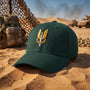 A green baseball cap with a gold and silver military-style SAS Who Dares Wins insignia on the front, resting on a sandy desert background with camouflage gear and military vehicles in the distance.