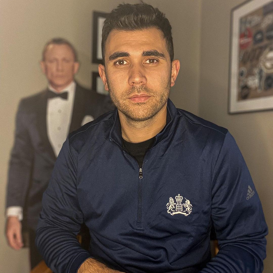 Luke from Bonds Apartment in a James Bond Adidas 1/4 Zip Pullover sitting in a room with James Bond wearing a Tux in the background.