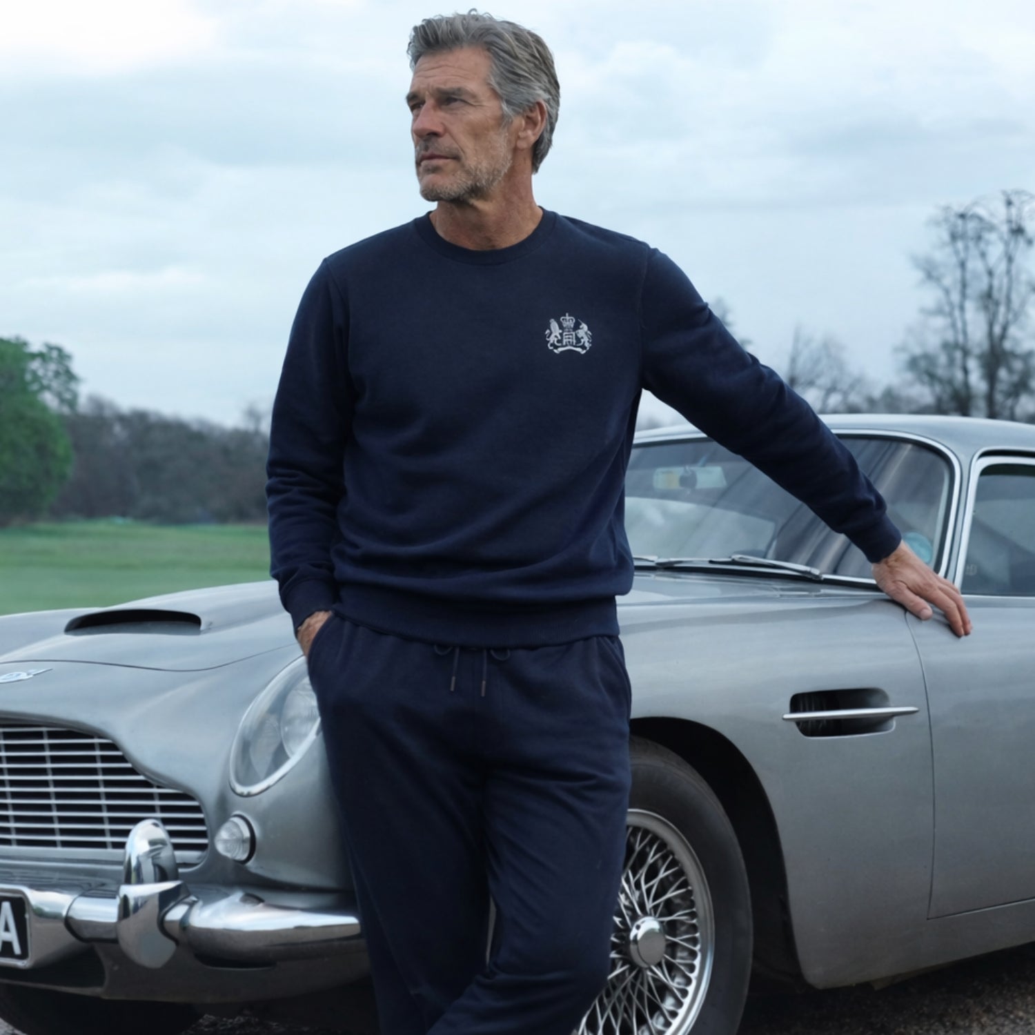 Man in James Bond Sweatshirt standing next to a classic Aston Martin DB5 Silver Birch with a cloudy sky background