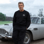 Man in James Bond Sweatshirt standing next to a classic Aston Martin DB5 Silver Birch with a cloudy sky background