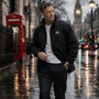Man walking on a rain-soaked street with iconic London landmarks in the background wearing a Black James Bond Windbreaker
