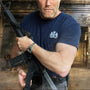 Man holding a rifle in an urban setting with a blurred background wearing a James Bond T-Shirt