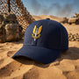 A navy blue baseball cap with a gold and silver military-style SAS Who Dares Wins insignia on the front, resting on a sandy desert background with camouflage gear and military vehicles in the distance.