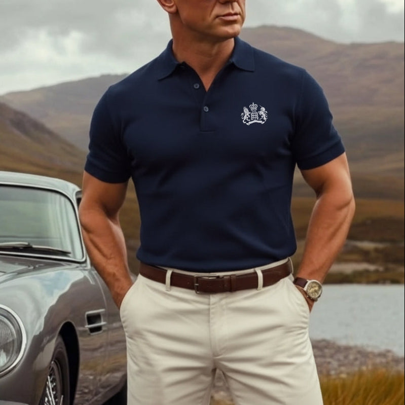 James Bond in a navy polo shirt standing next to a Aston Martin DB5 with mountains in the background