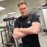 Man in a gym wearing a black t-shirt with a logo, arms crossed.