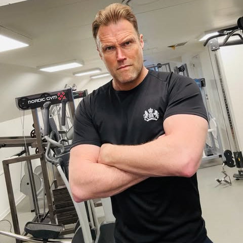 Man in a gym wearing a black t-shirt with a logo, arms crossed.