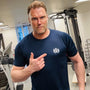 Man in a gym wearing a blue t-shirt with a logo, pointing at the camera.