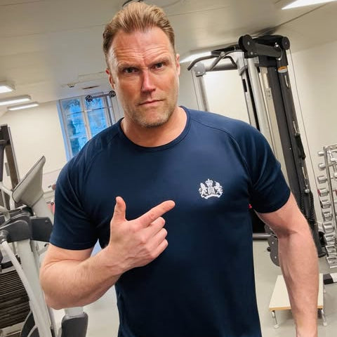 Man in a gym wearing a blue t-shirt with a logo, pointing at the camera.