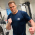 Man in a gym wearing a blue t-shirt with a logo, flexing his muscles.