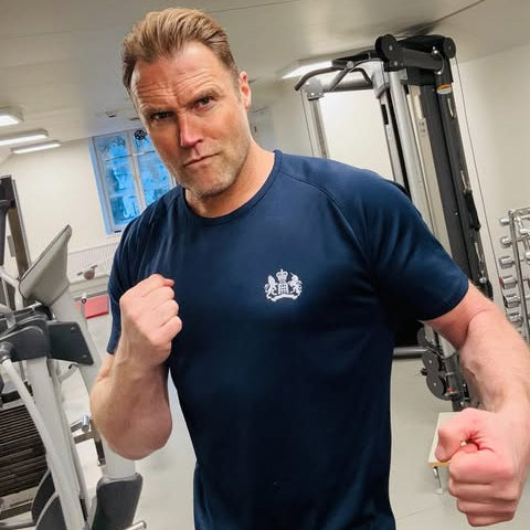Man in a gym wearing a blue t-shirt with a logo, flexing his muscles.