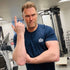 Man in a gym wearing a blue t-shirt with a logo, posing with a hand gesture.