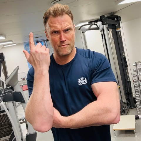 Man in a gym wearing a blue t-shirt with a logo, posing with a hand gesture.