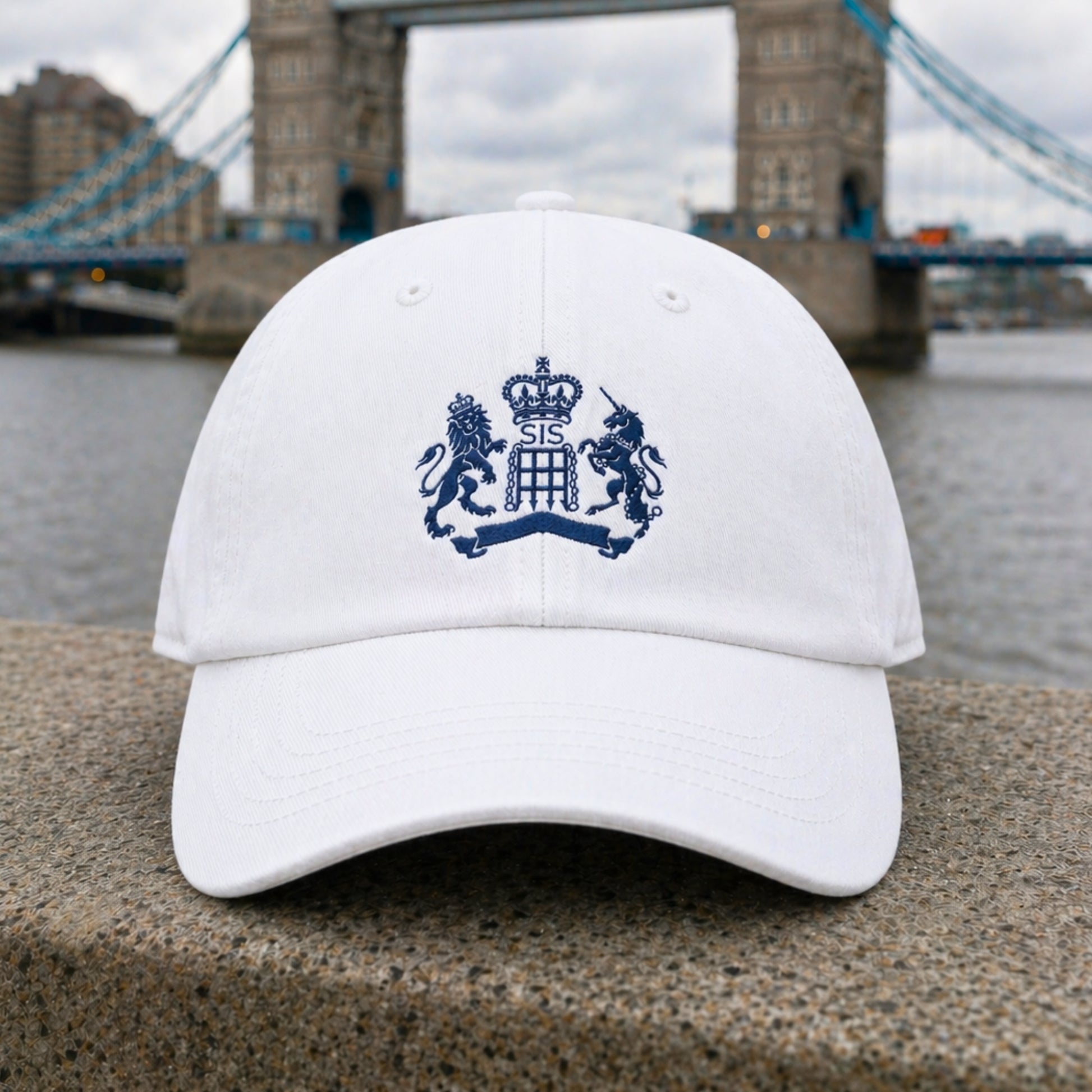 White James Bond Hat with Embroidered S.I.S. Logo from Skyfall on a stone ledge with a bridge and river in the background